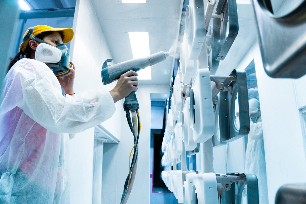 Powder coating of metal parts. A woman in a protective suit sprays white powder paint from a gun on metal products
